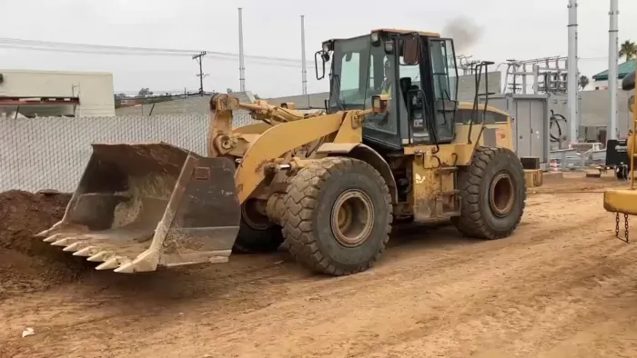 A complete view of a yellow 1999 Cat 950G wheel loader driving on a dirt road within a city setting.