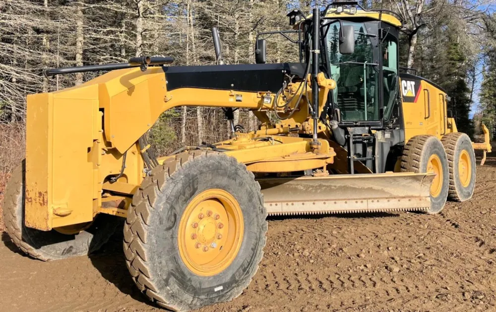 A yellow Caterpillar motor grader working on a dirt surface, with trees visible behind it.