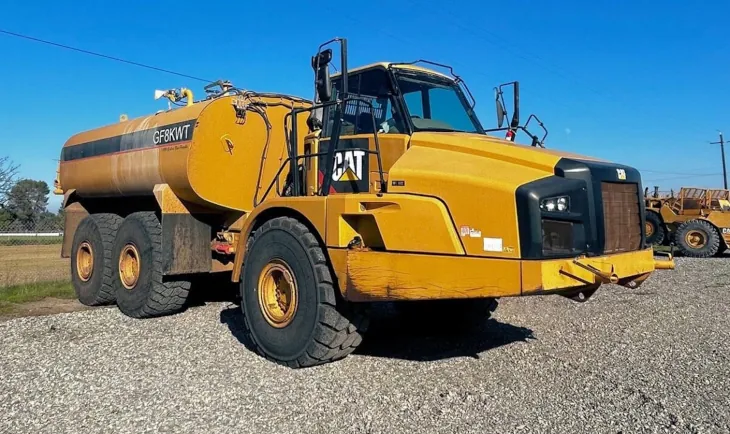 A detailed view of a yellow 2012 Cat 740B Water truck situated on a gravel lot beneath a clear blue sky.