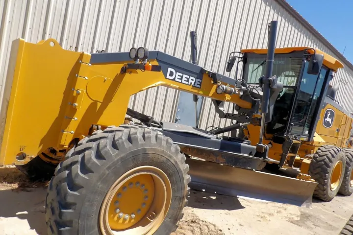 A big yellow 2022 John Deere 672GP motor grader stationed on a dirt lot, with a corrugated metal wall behind it.