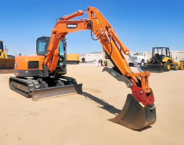 A compact orange 2023 Doosan excavator positioned on a dirt lot, with additional heavy equipment visible in the background.