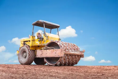 A massive yellow spiked compactor is stationed on a dirt lot beneath a clear blue sky.