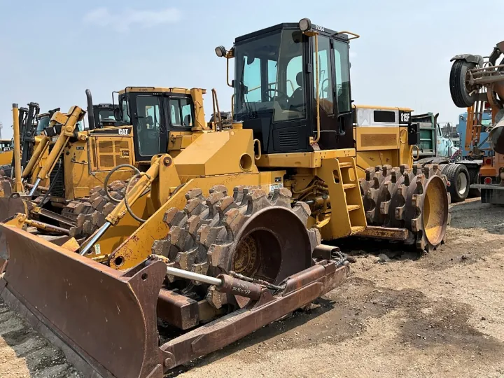 A detailed view of a yellow 2004 Cat 815F compactor, featuring a prominent front blade and oversized, spiked tires designed for soil compaction, parked on a dirt lot among various other heavy machines.