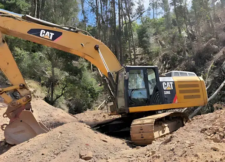 A yellow 2013 Cat 336E excavator is parked among several dirt mounds, with a forested area in the background.