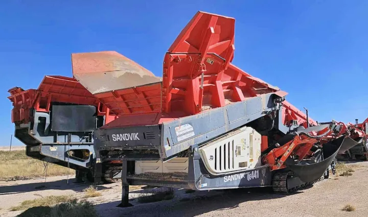 A 2018 Sandvik QE441 Scalper, a large aggregate machine with a red upper section, parked among other equipment in a lot.