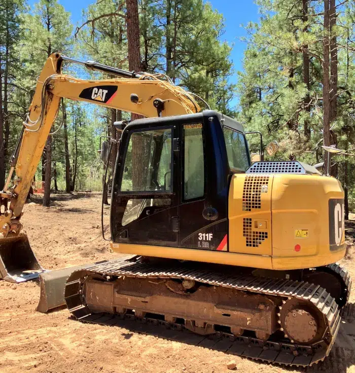 A 2014 Caterpillar 311FLRR excavator machine in yellow is parked among pine trees in the forest.